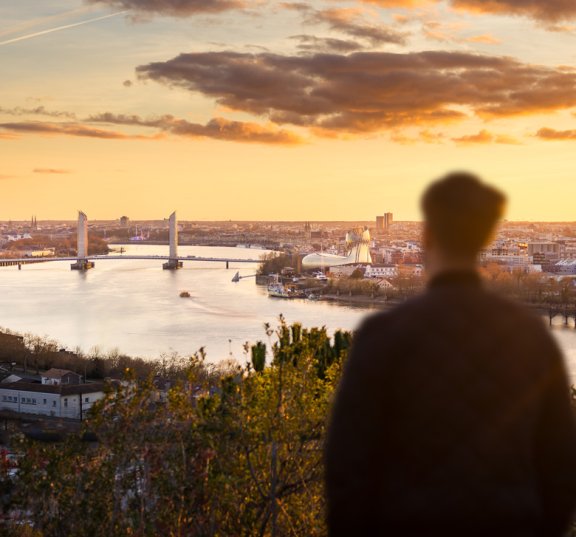 Une personne de dos regarde la Garonne, le pont Chaban-Delmas et la cité du vin, à l'aube, depuis le parc de l'Hermitage à Lormont. 